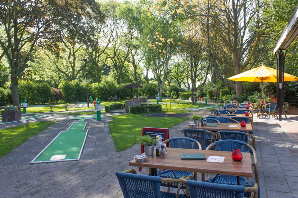 Outdoor café with blue wicker chairs and wooden tables beside a mini-golf course, surrounded by lush green trees and park scenery.