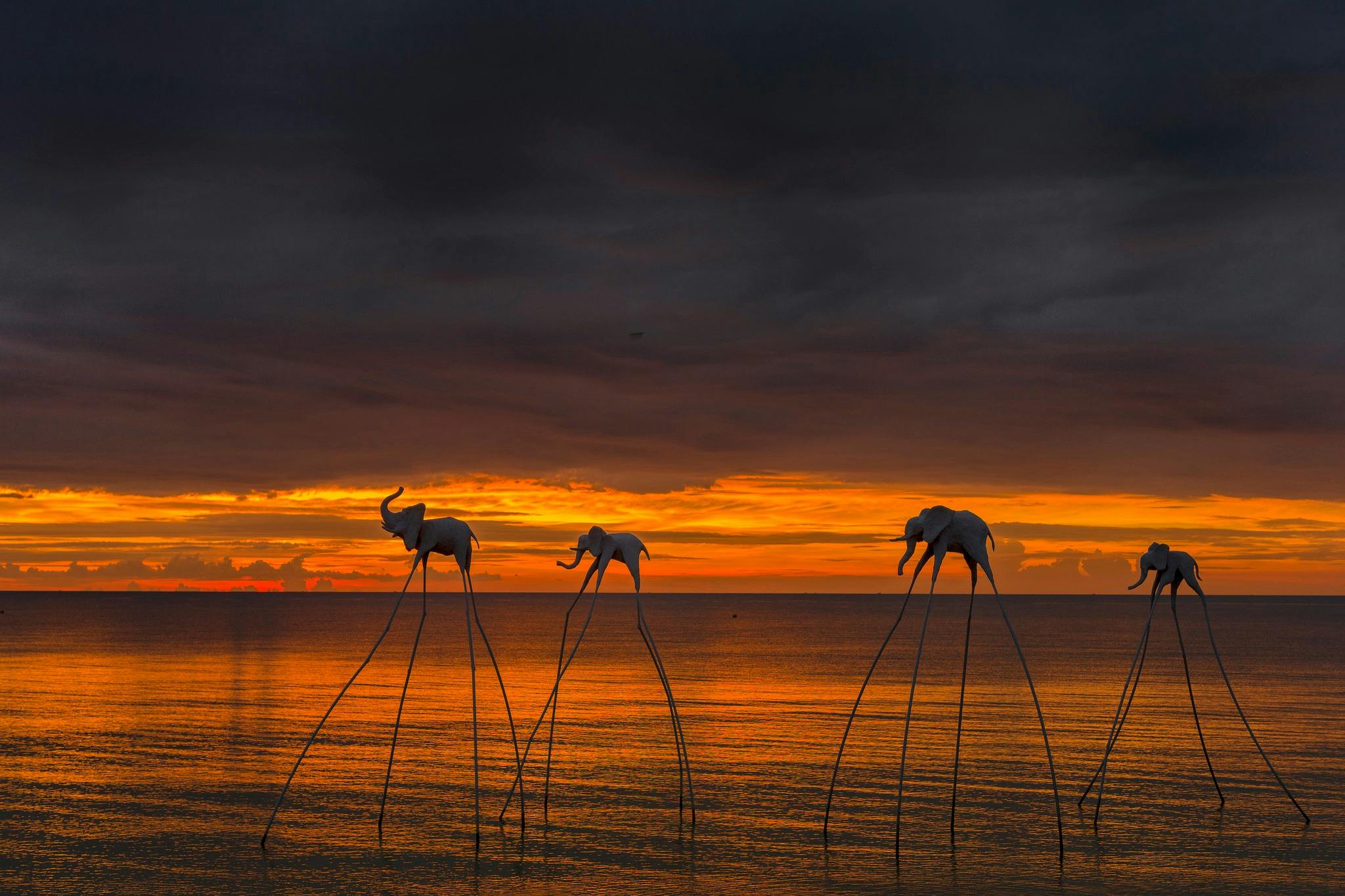 Three surreal elephant sculptures stand on tall, thin legs in shallow water at sunset under a dramatic, cloudy sky.