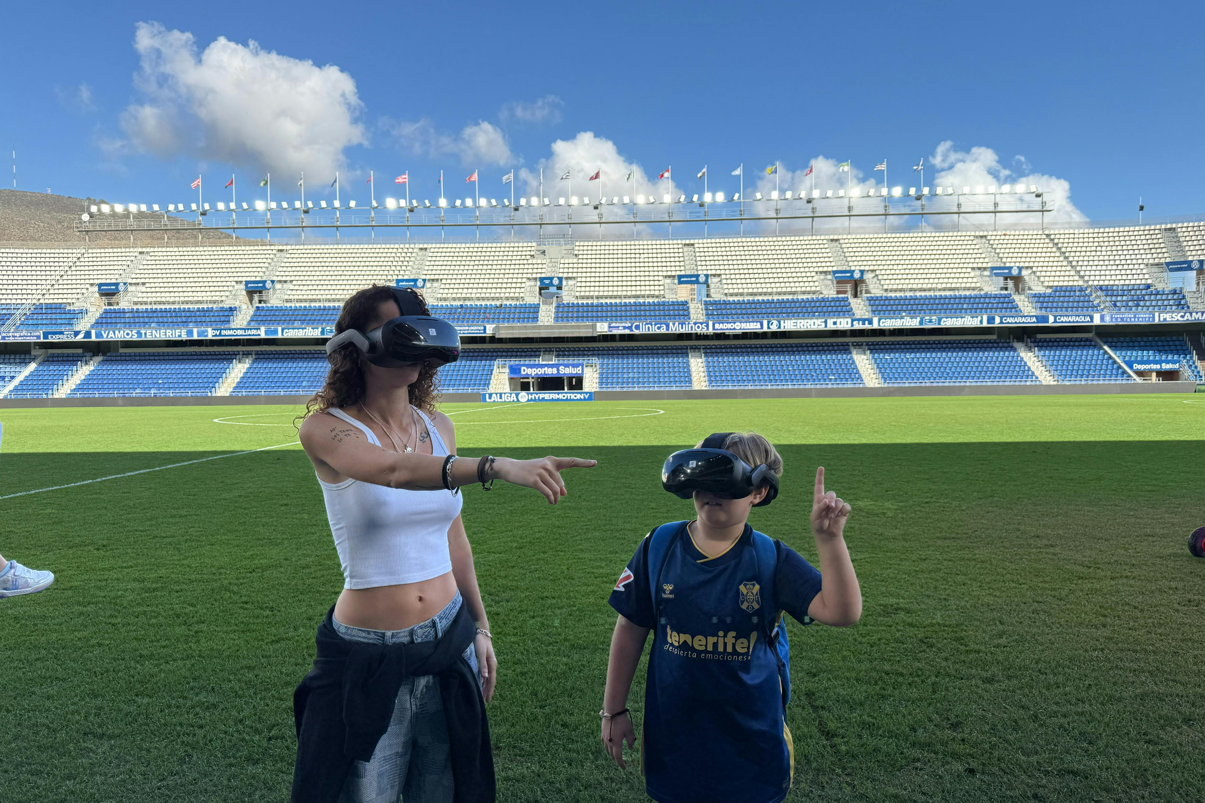 Dos persones amb auriculars de realitat virtual estan parades en un camp de futbol. Els seients de l'estadi i el cel brillant són al fons.