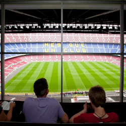 Fyra personer observerar en stor, tom fotbollsstadion från en hög utsiktspunkt, med säten som stavar "mes QUE FN-KLUBB".