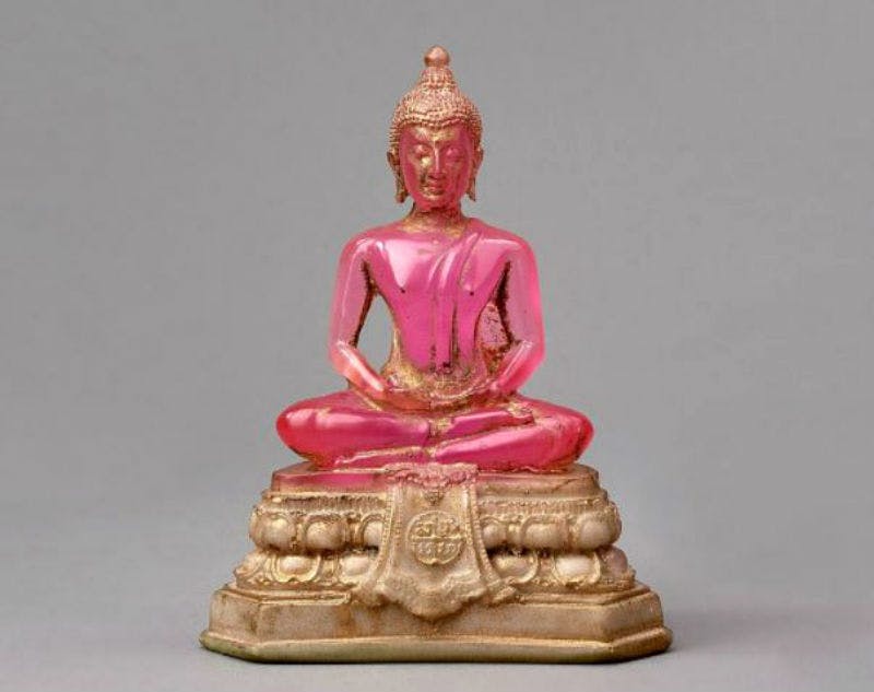 A small, pink, seated Buddha statue on an ornate, golden pedestal against a plain gray background.