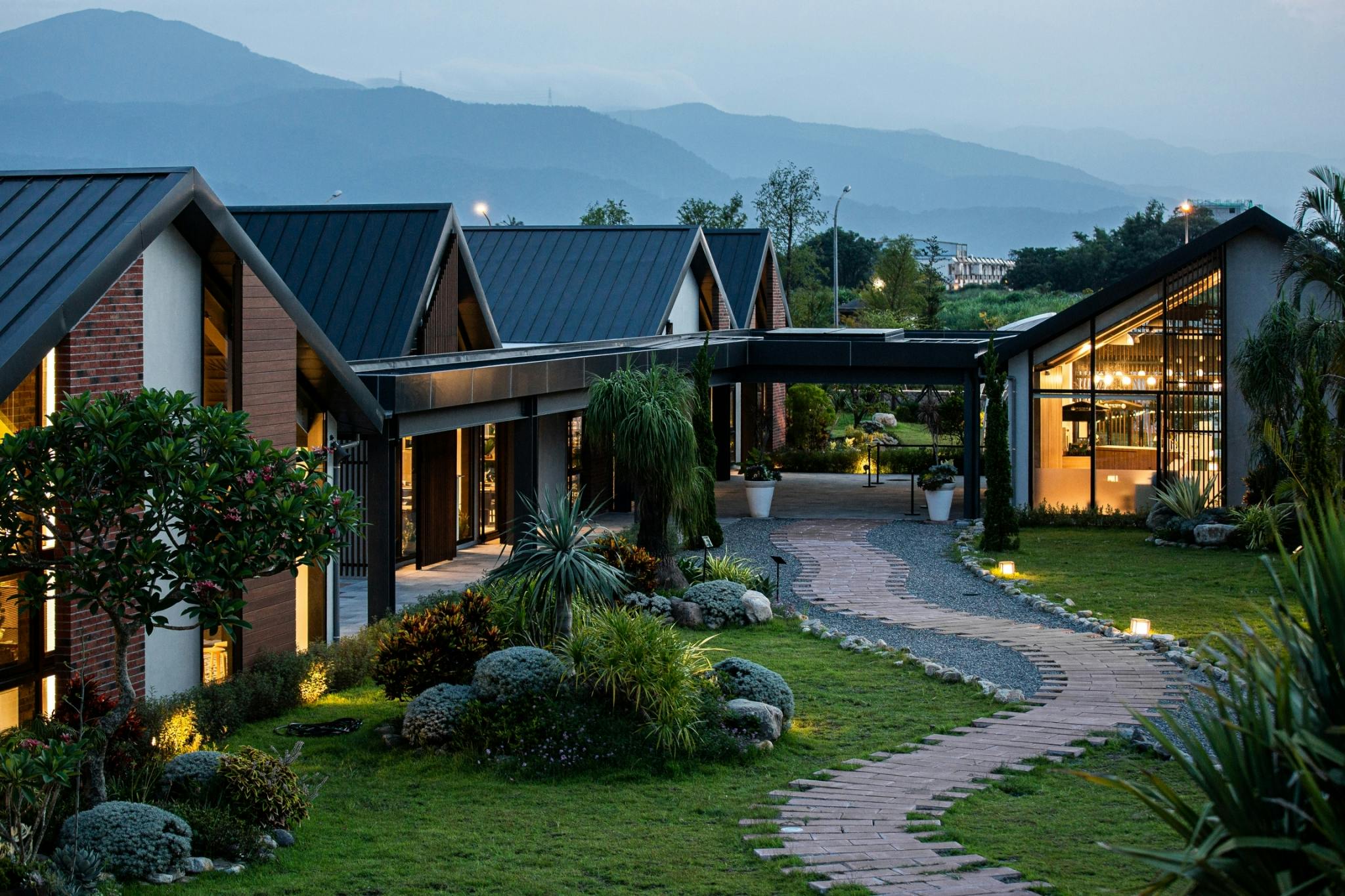 A modern complex of buildings with slanted roofs surrounded by landscaped gardens and mountains in the background.