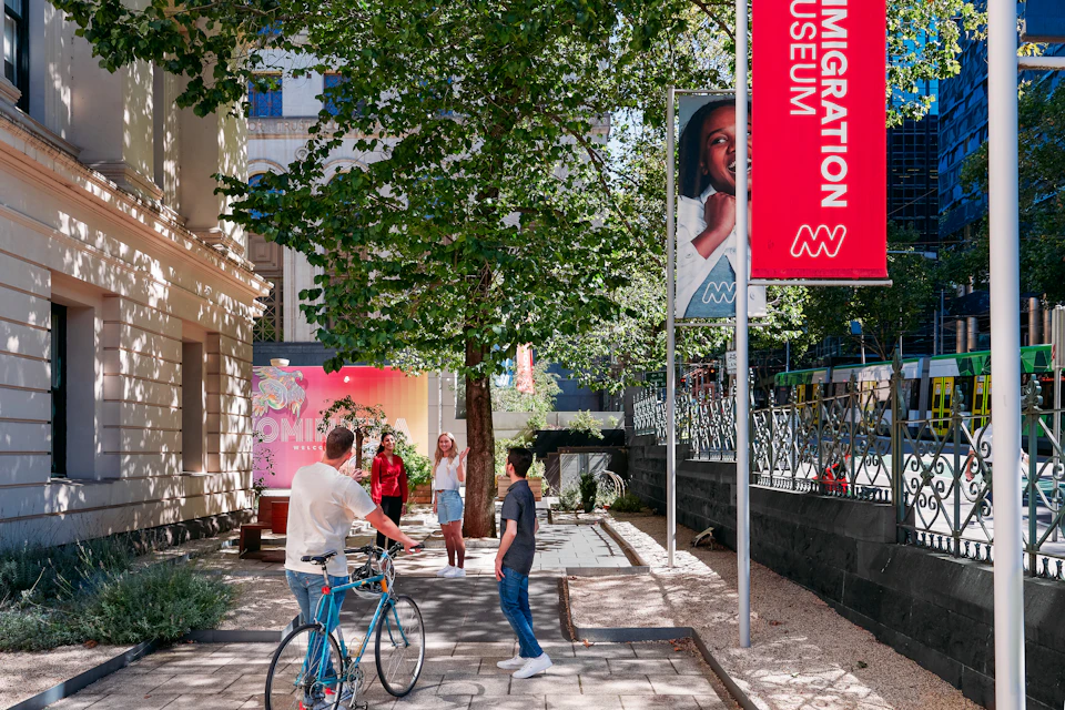 People walking and cycling near a tree-lined path outside the Immigration Museum, next to a red and white museum banner.