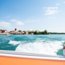 View from a boat of a small town, with wake on water, under a clear blue sky. Nearby land has buildings and greenery.