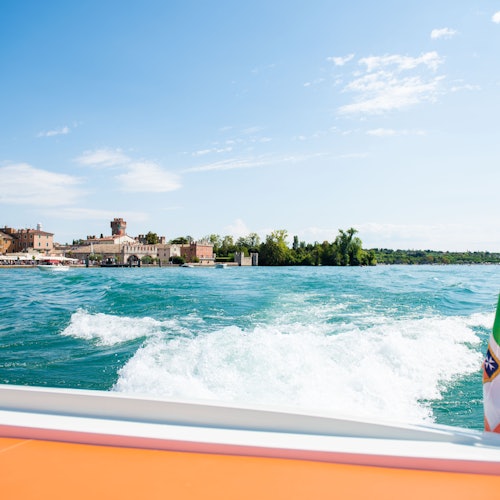 View from a boat of a small town, with wake on water, under a clear blue sky. Nearby land has buildings and greenery.