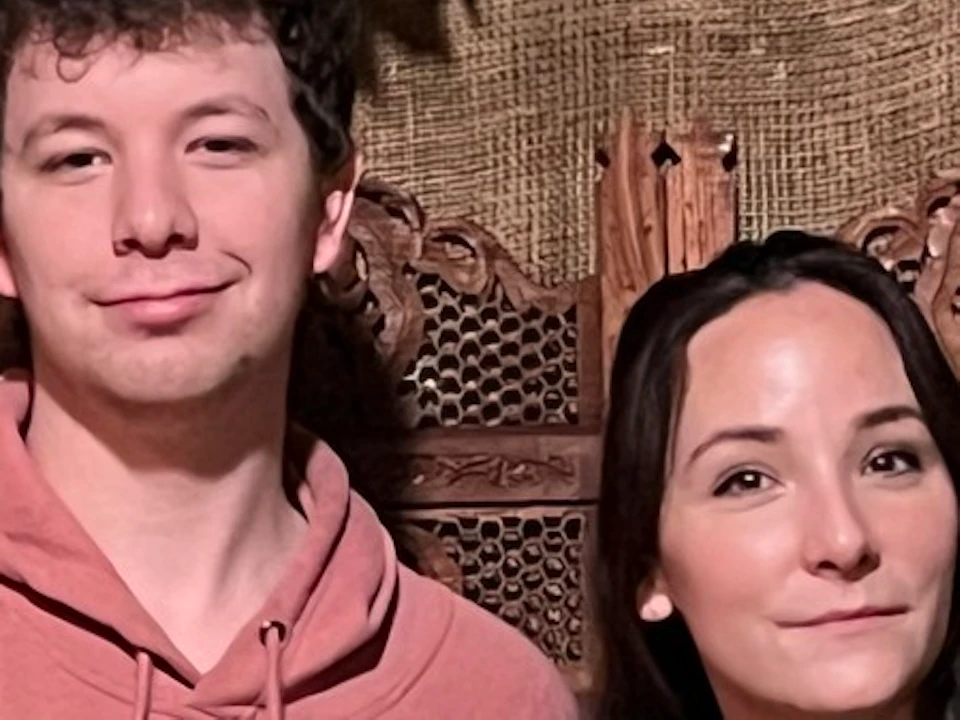 Two people standing with parrots perched on their arms in front of a wooden screen, surrounded by green foliage.