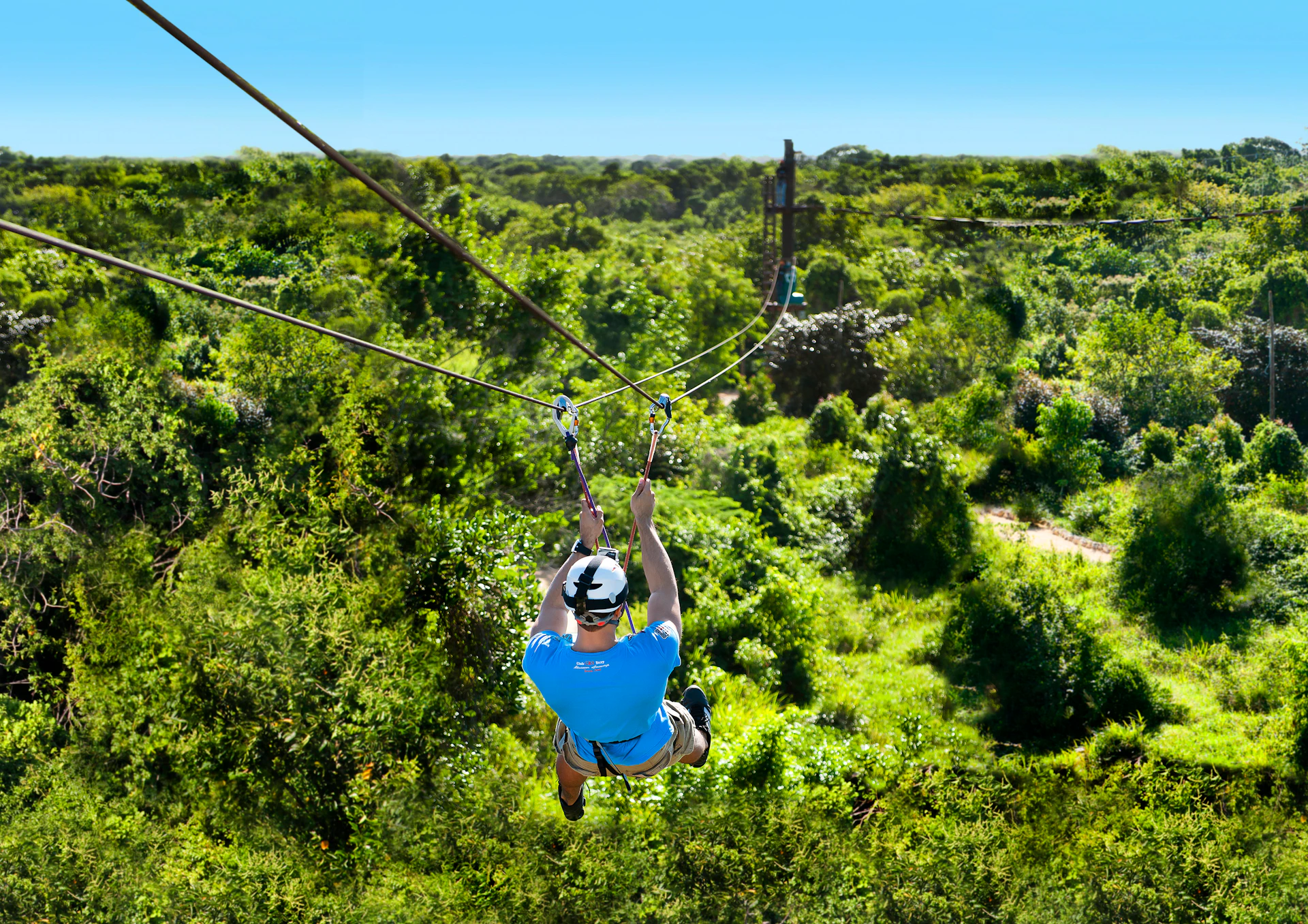 Person ziplining over lush green forest, wearing a blue shirt, shorts, and helmet. Sky is clear with bright sunlight.