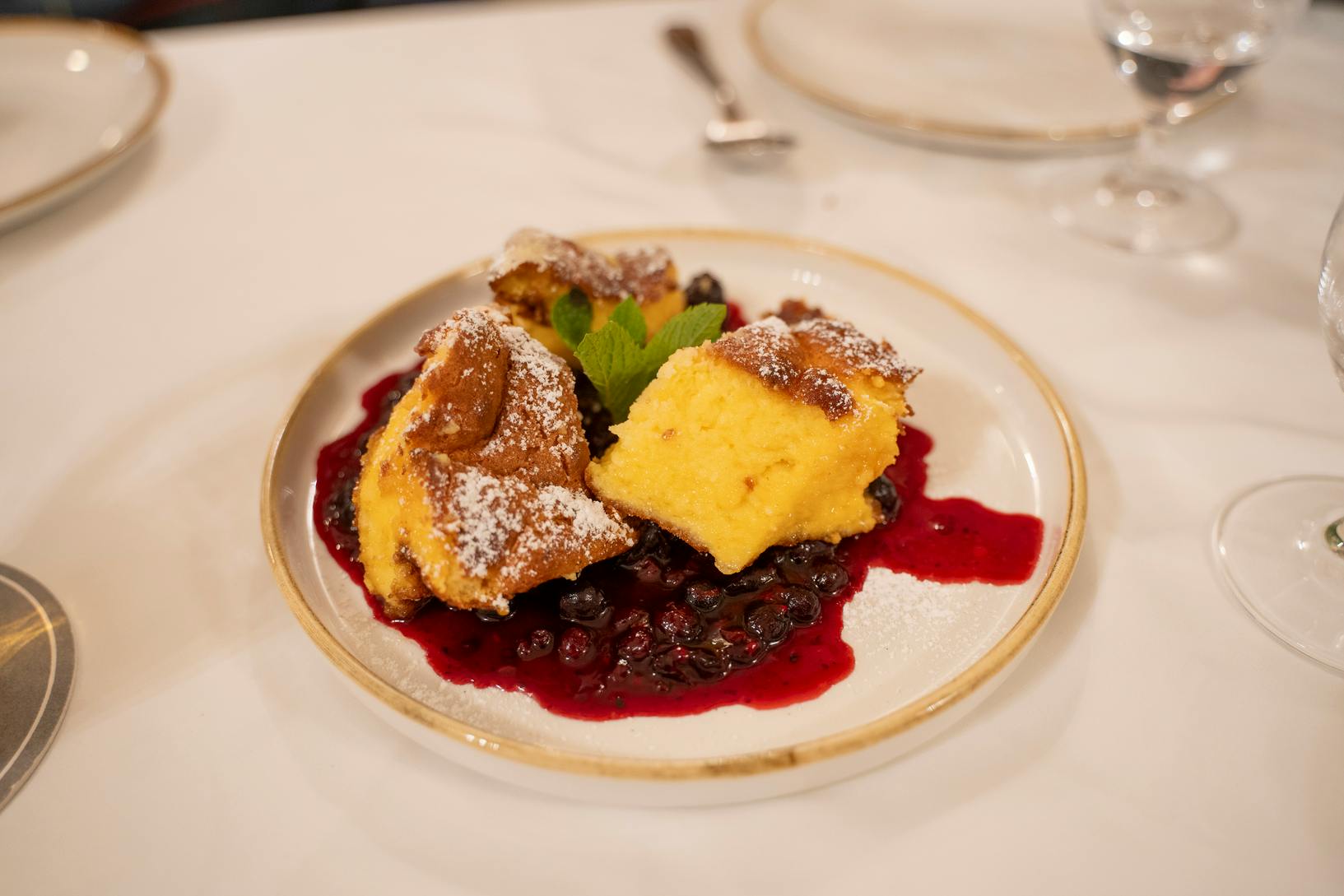 Um prato com pedaços de massa polvilhada com açúcar em pó servido numa cama de compota de frutos silvestres, guarnecido com uma folha de hortelã.