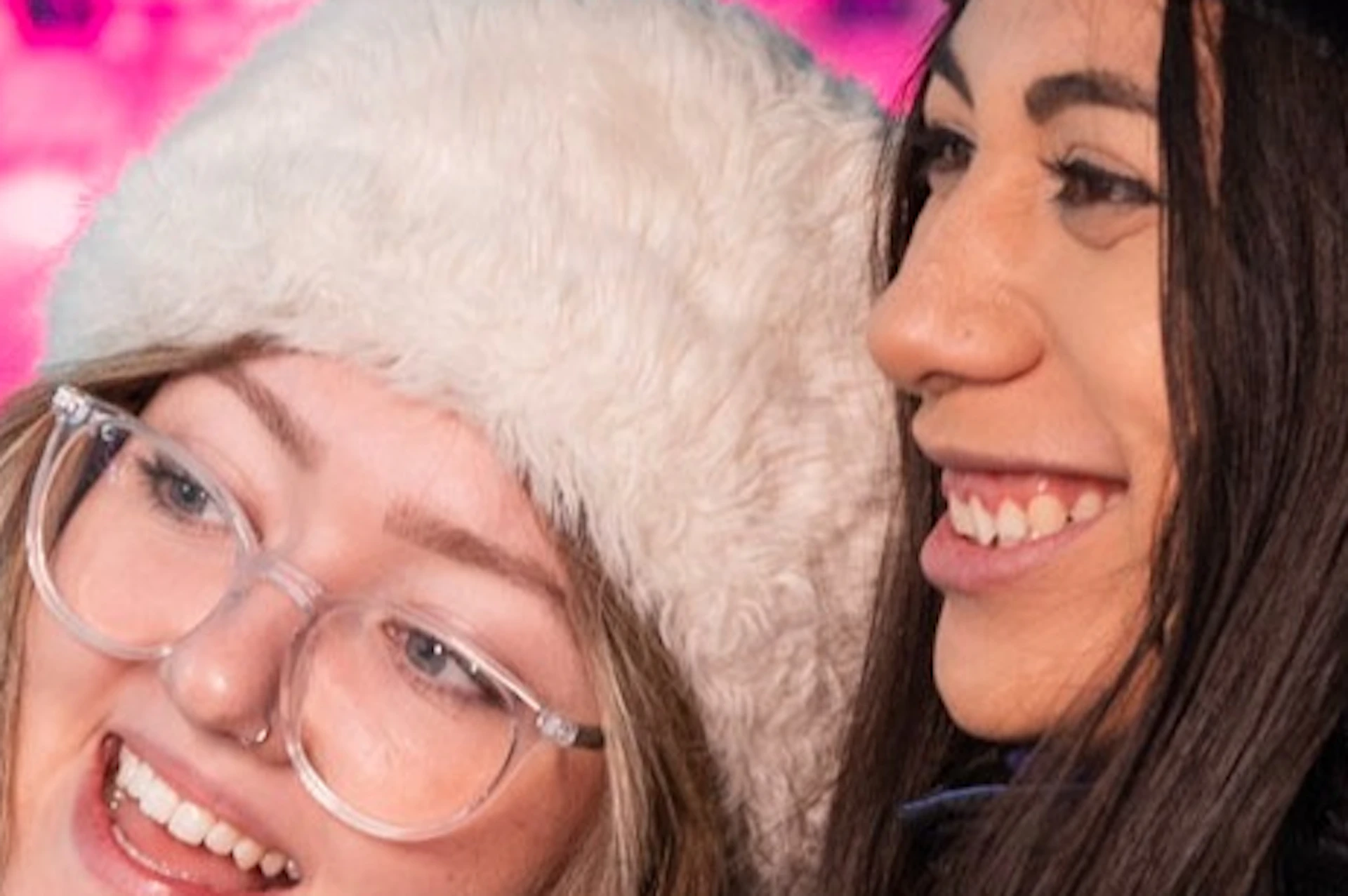 Two people wearing winter hats and coats smile in an illuminated environment with pink and blue lighting.