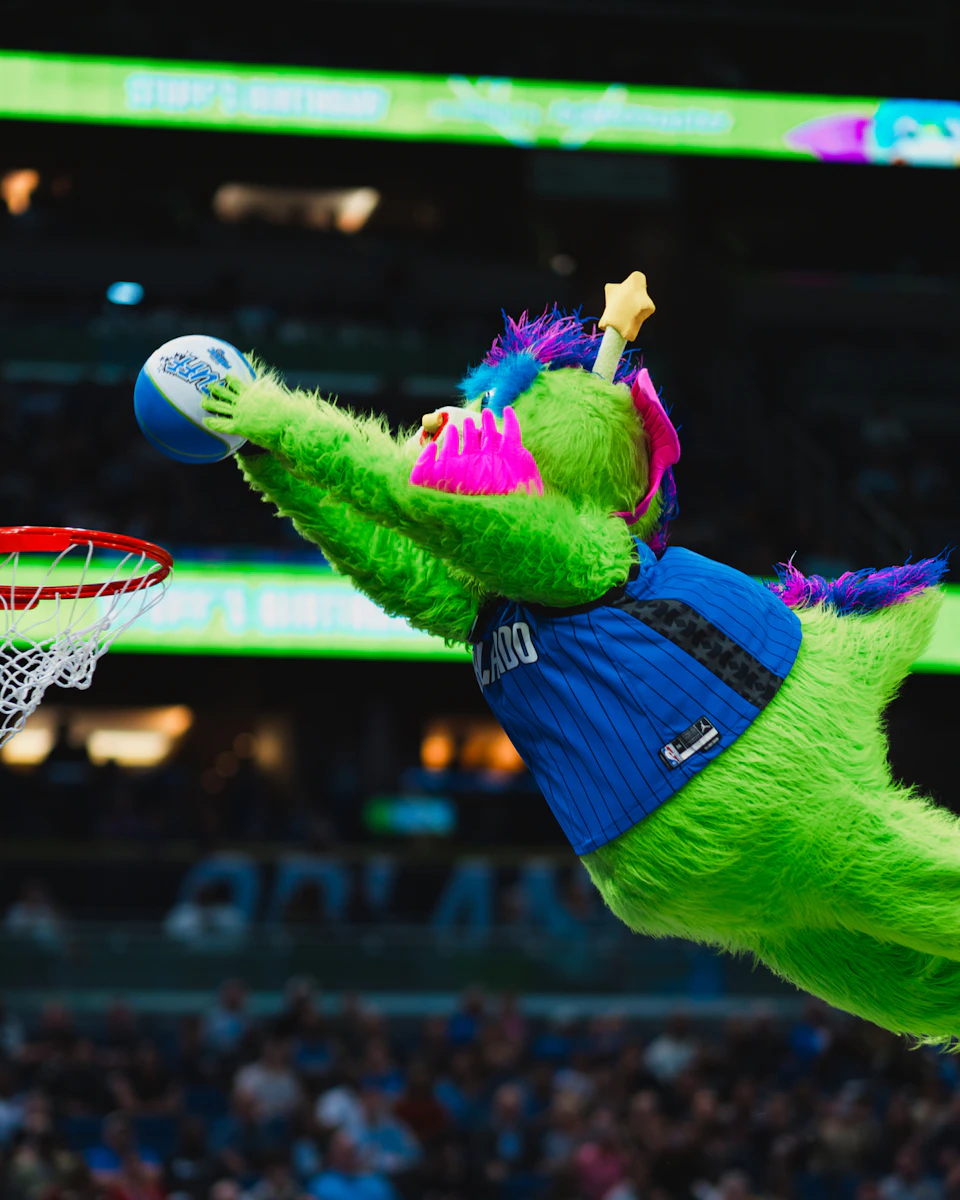 A green mascot in a blue jersey jumps and dunks a basketball in a sports arena.