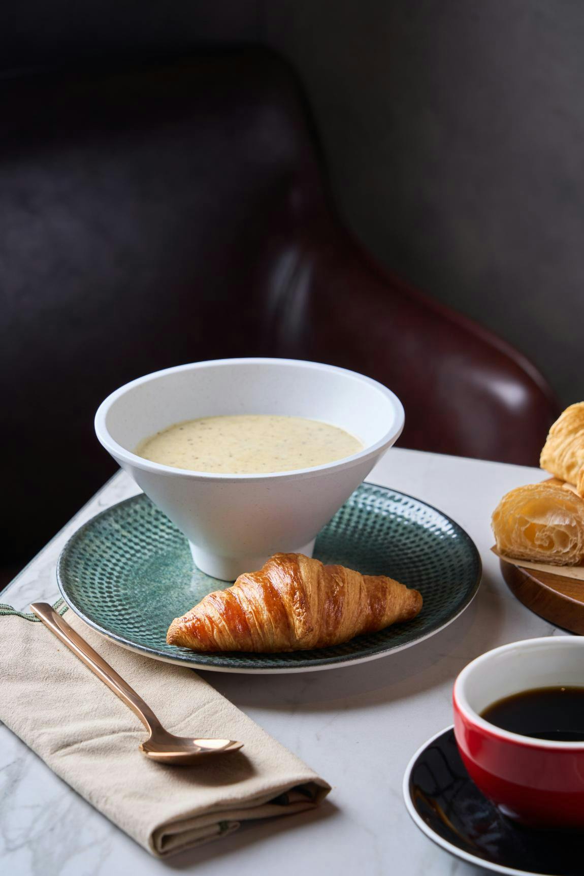 A bowl of soup on a green plate with a croissant, a napkin, and pastries on a table.