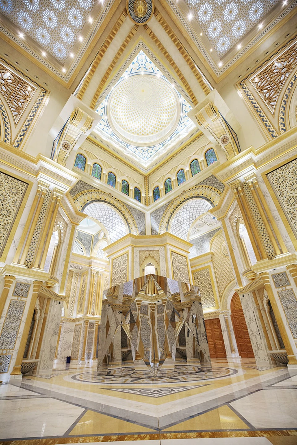 An opulent interior features ornate arches, columns, intricate patterns, a dome ceiling, and large mirrors reflecting the space.