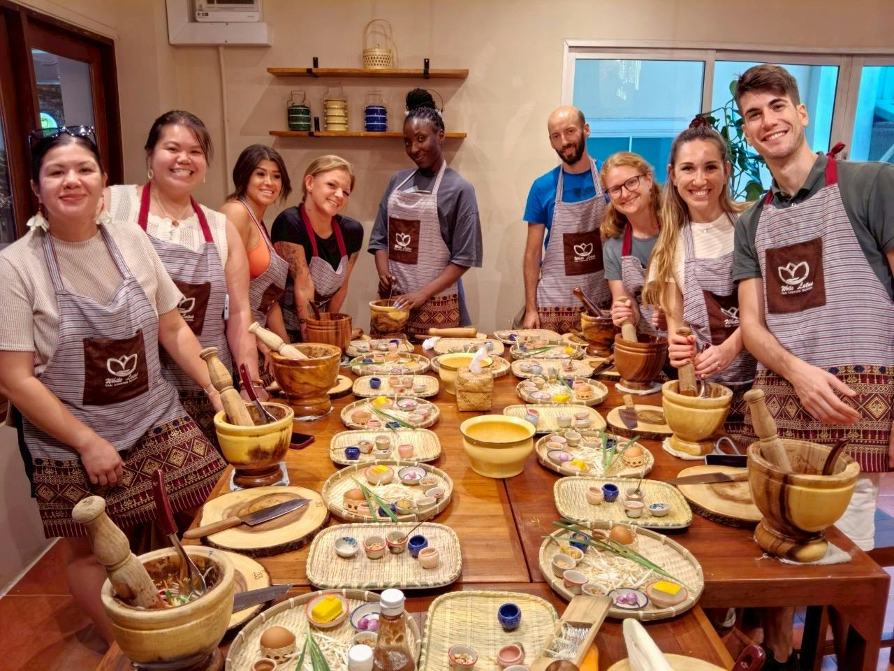 A group of people in aprons smiling around a table set with cooking ingredients and utensils in a cooking class setting.