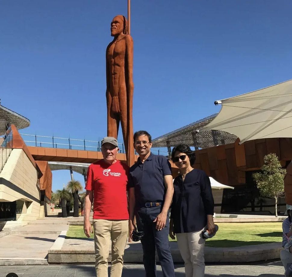 三个人站在一座高大的铁锈色人物雕像前。 背景是现代建筑和结构。