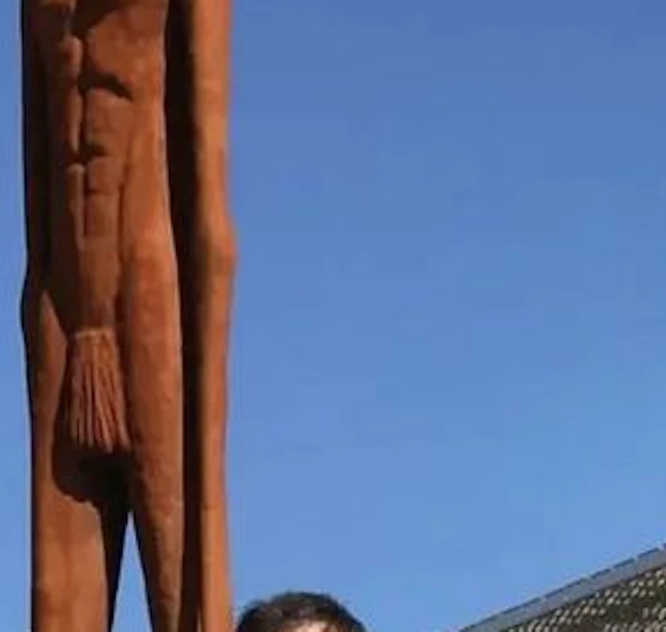 Three people standing in front of a tall, rust-colored statue of a human figure. Modern buildings and structures are in the background.