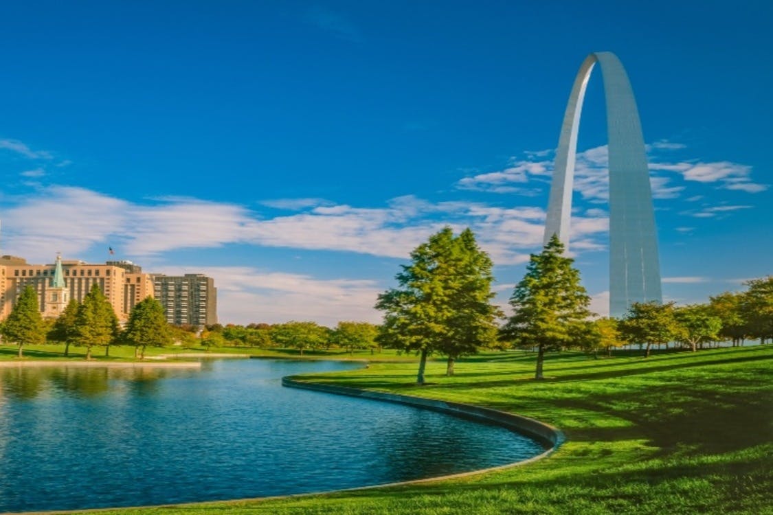 Μια μεγάλη καμάρα στέκεται κοντά σε μια καμπύλη όχθη ποταμού που περιβάλλεται από καταπράσινο γρασίδι και δέντρα, με κτίρια στο παρασκήνιο κάτω από ένα γαλάζιο ουρανό.