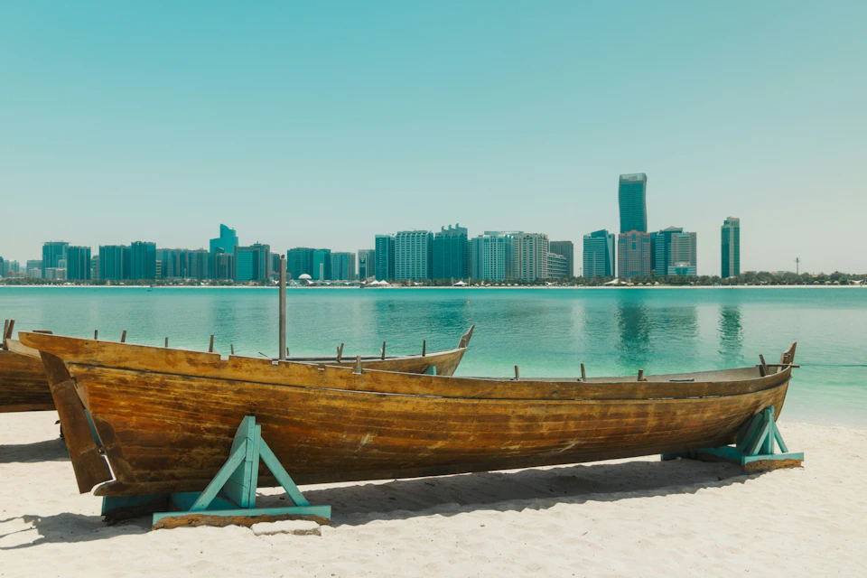 A wooden boat on a sandy beach with a city skyline and water in the background under a clear sky.