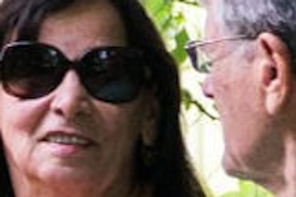 Two people sitting on a wooden bench in a lush green garden, surrounded by plants and trees, having a conversation.