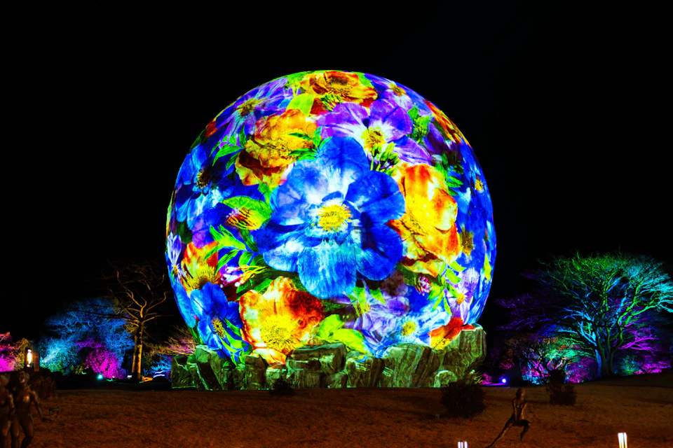 A spherical structure illuminated with vibrant floral projections under a night sky, surrounded by trees and lights.