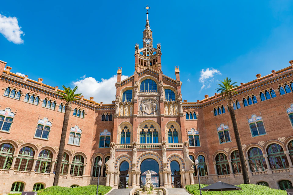 Ornate historical building facade with intricate arches, decorative windows, and a tall central spire, flanked by palm trees. Clear blue sky.