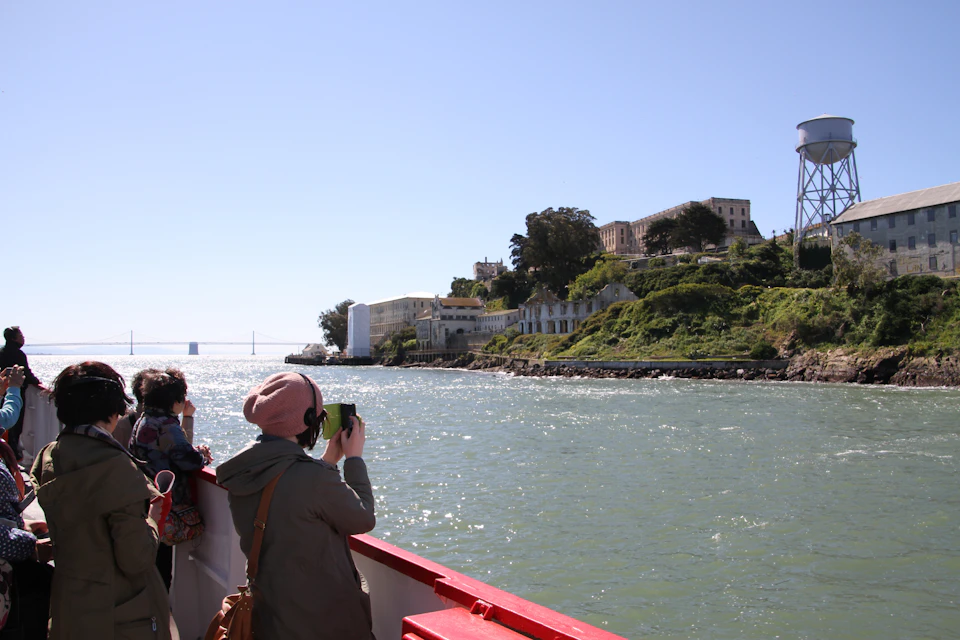 Two people on a boat taking photos of a distant island with historic buildings on a sunny day.