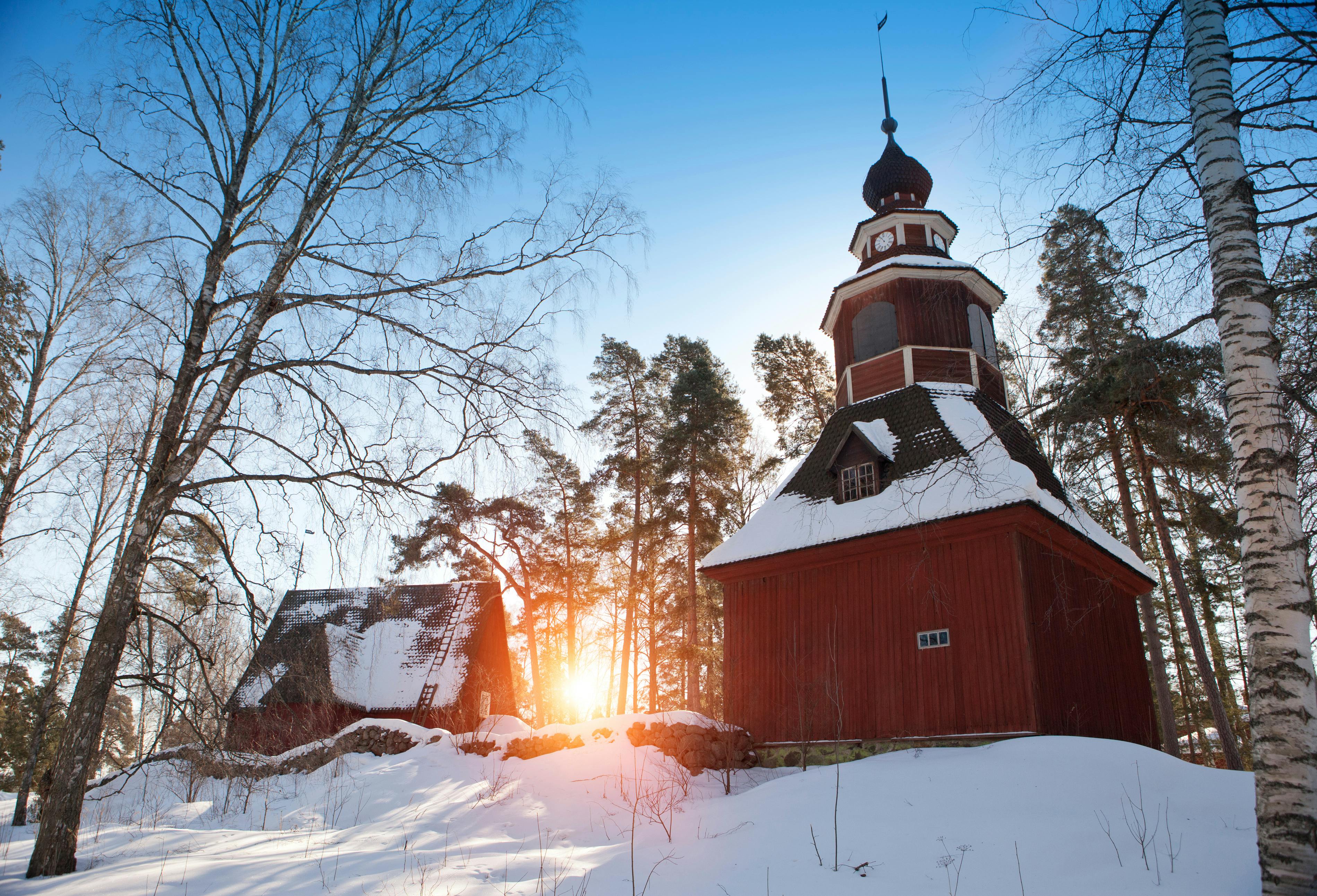 Ett snöigt landskap med en röd träkyrka och en annan byggnad, omgiven av nakna träd, mot en kuliss av en nedgående sol.