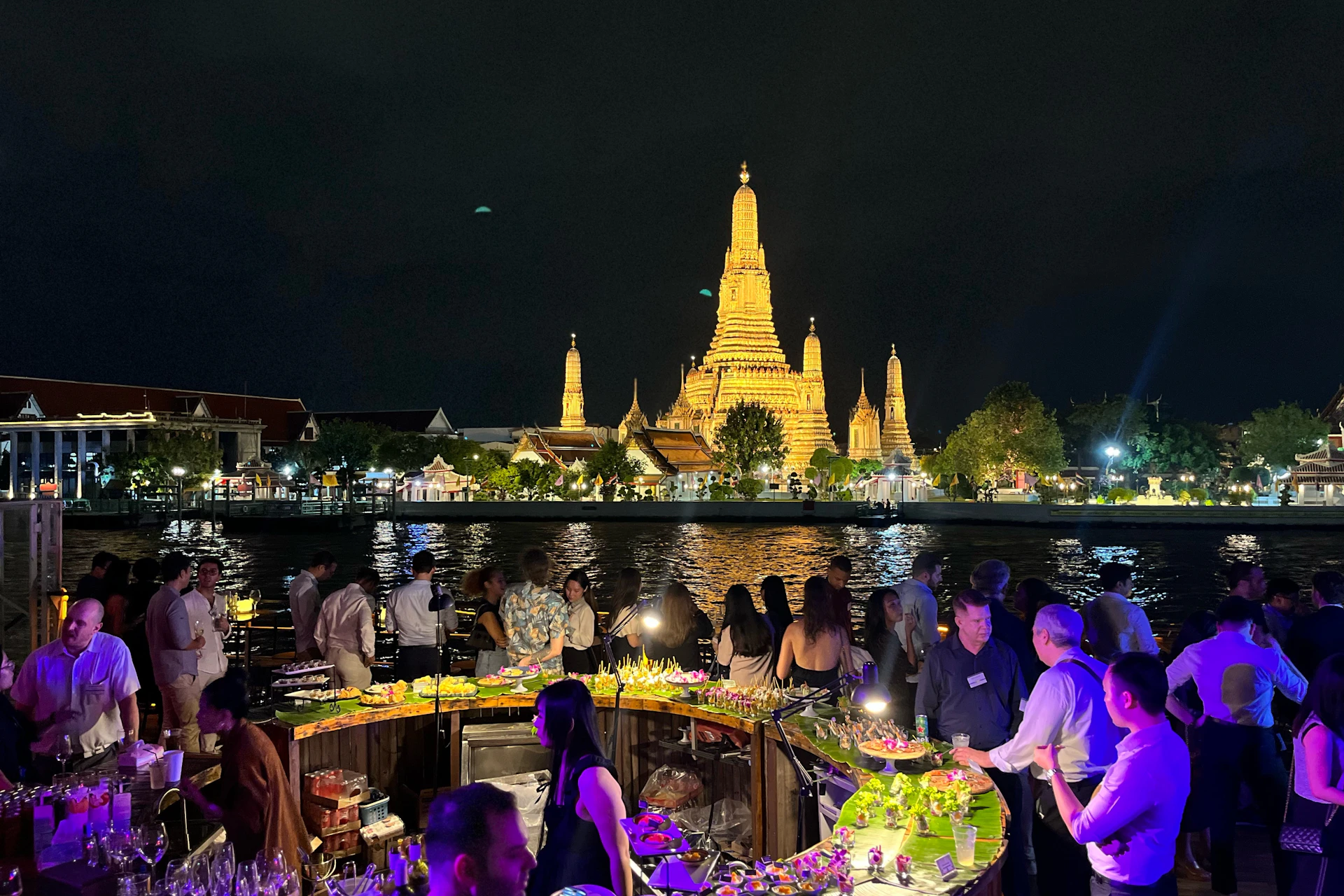 People socializing at an outdoor evening event near a river, with a brightly lit temple in the background.