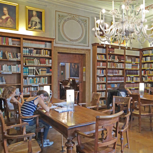 Folk studerar vid träbord i ett historiskt bibliotek med bokhyllor, inramade porträtt och en ljuskrona.
