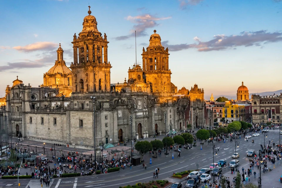 A historic cathedral is illuminated by the setting sun, with a busy street and people gathered below, alongside buildings and trees.