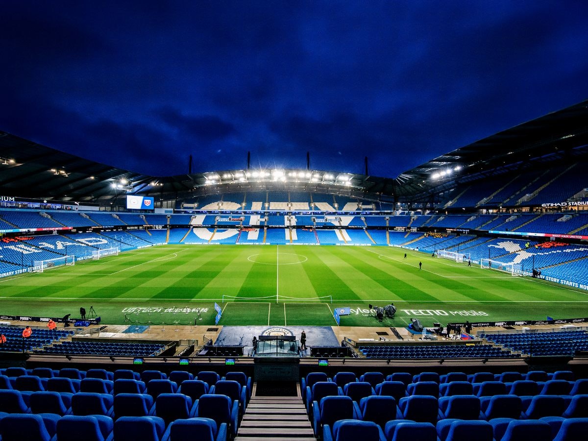 Pusty stadion piłkarski z niebieskimi siedzeniami i zielonym polem, oświetlonym w nocy. Widoczne są dwie tablice wyników i słupki bramkowe.