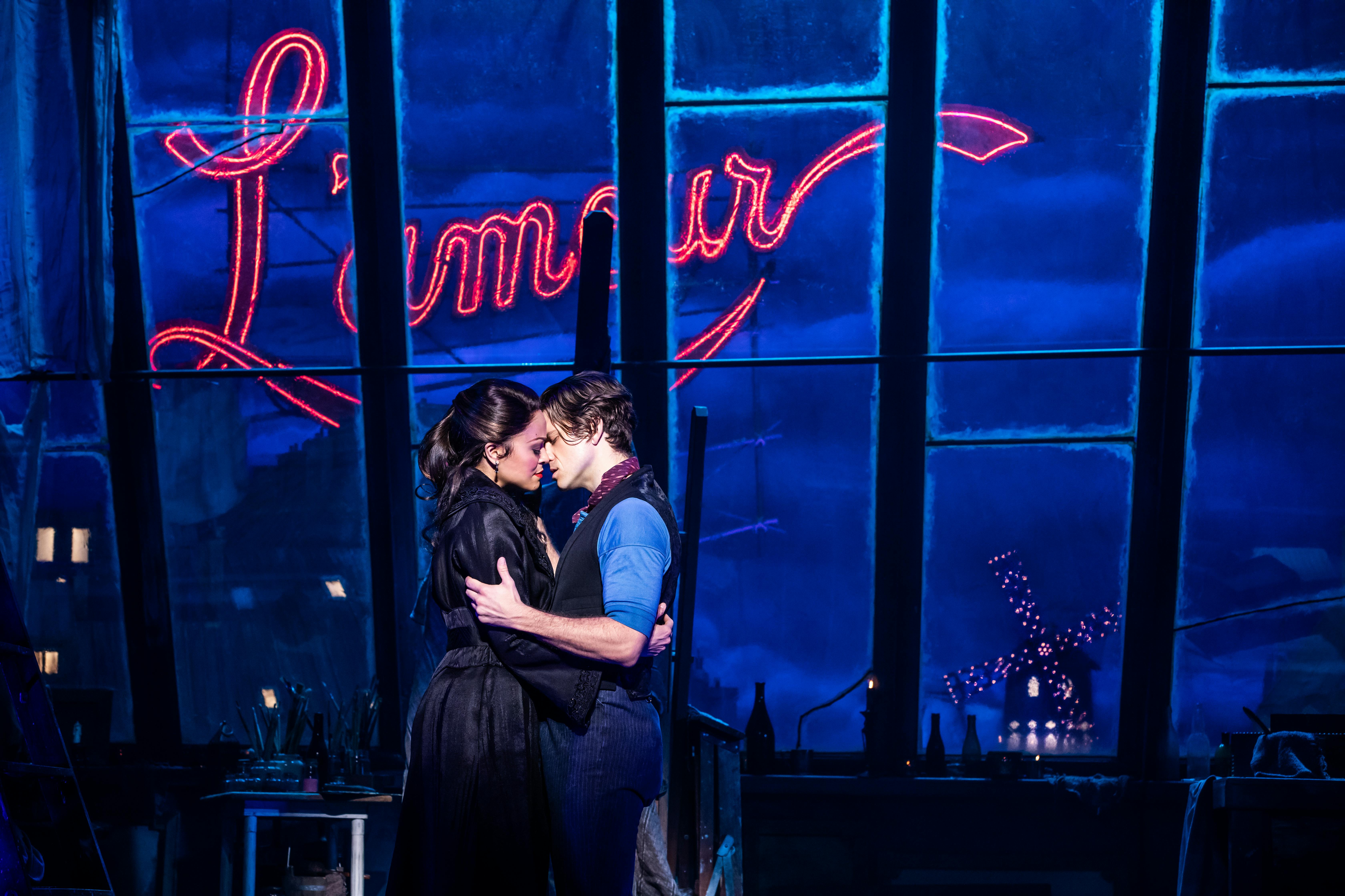 A couple embraces intimately under a neon “L’amour” sign in a dimly lit, blue-tinted environment.