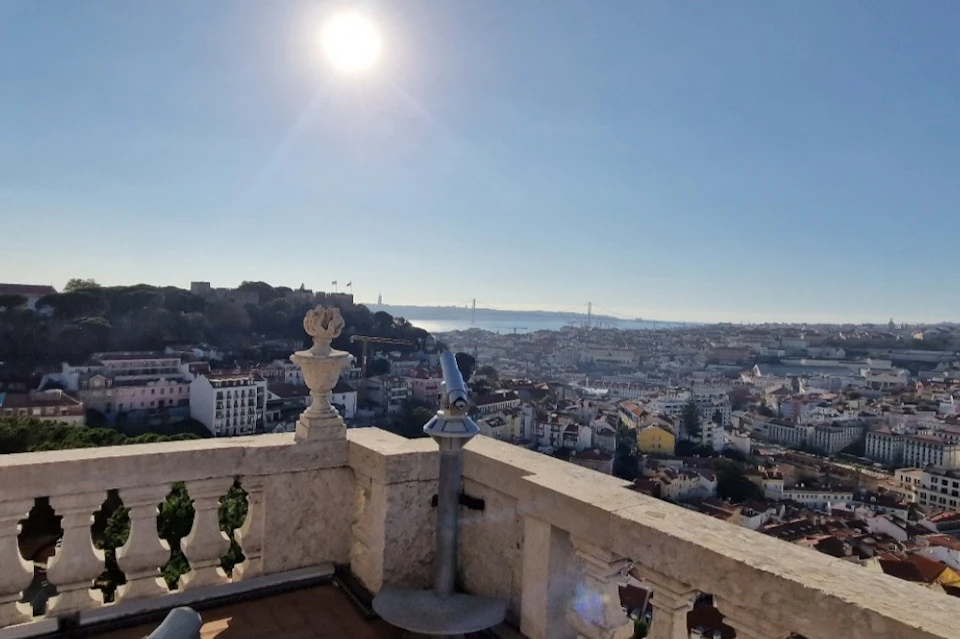 A scenic view of a city with rooftops, a bridge in the background, and a public binocular telescope on a stone balcony.