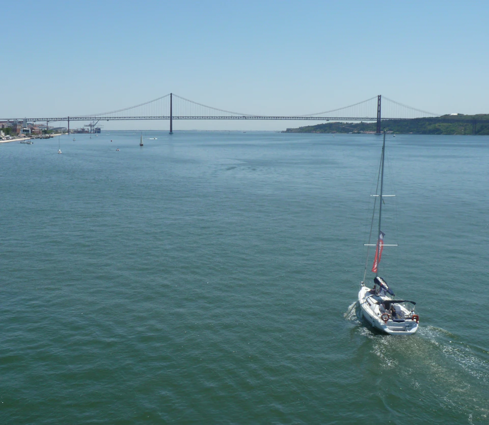 Sailboat moving on calm water towards a distant suspension bridge under a clear blue sky.