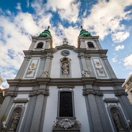 Vit barockkyrkofasad med två torn, gröna kupoler, statyer, reliefer och en klocka, under en delvis molnig blå himmel.
