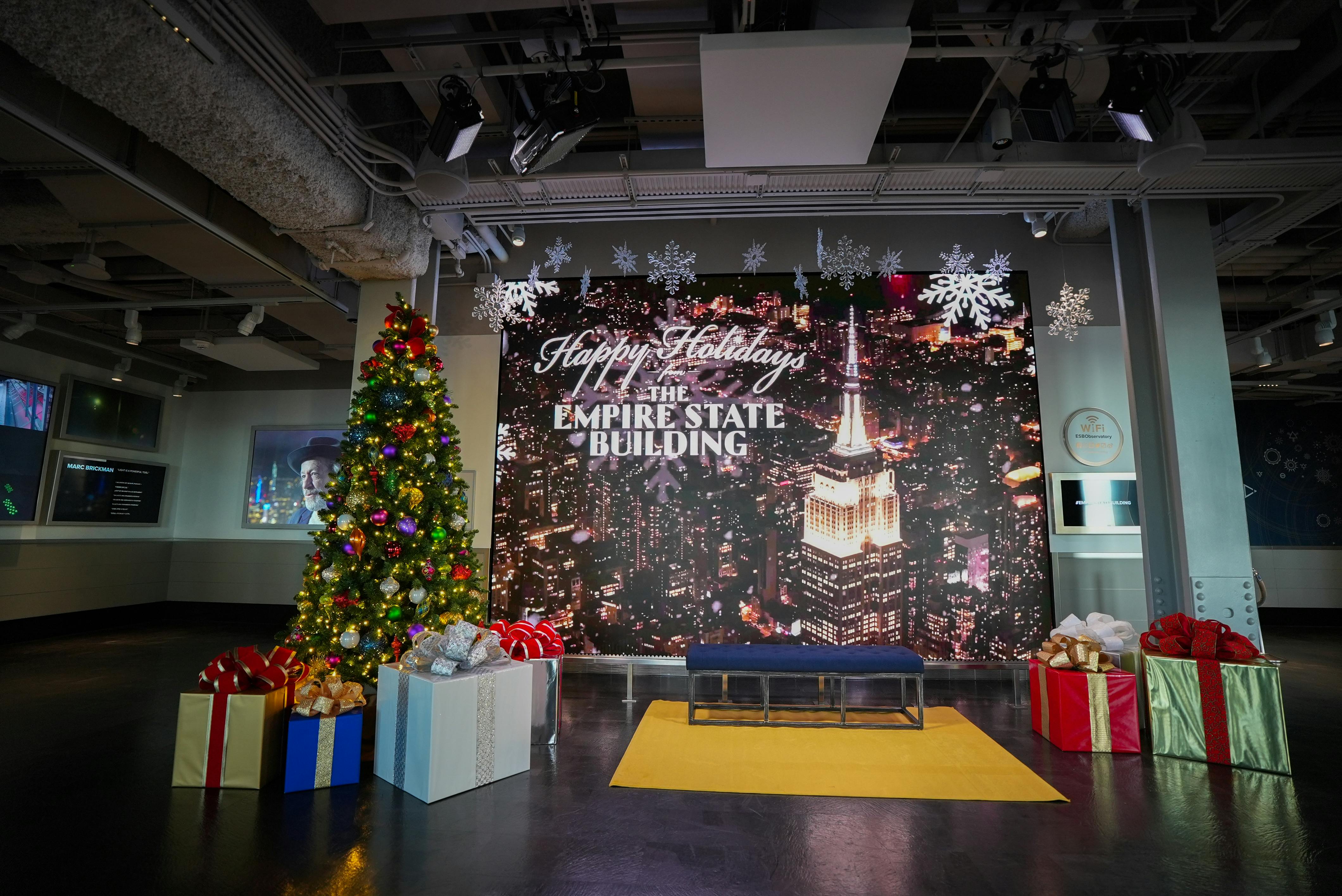 En festlig juleudstilling med et dekoreret juletræ, gaver og en baggrund af Empire State Building om aftenen.