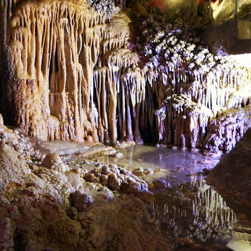 Ein Höhleninneres mit Stalaktiten- und Stalagmitenformationen, einem kleinen Wasserbecken, in dem sich das Licht spiegelt, und mit Mineralien bedeckten Felsen.