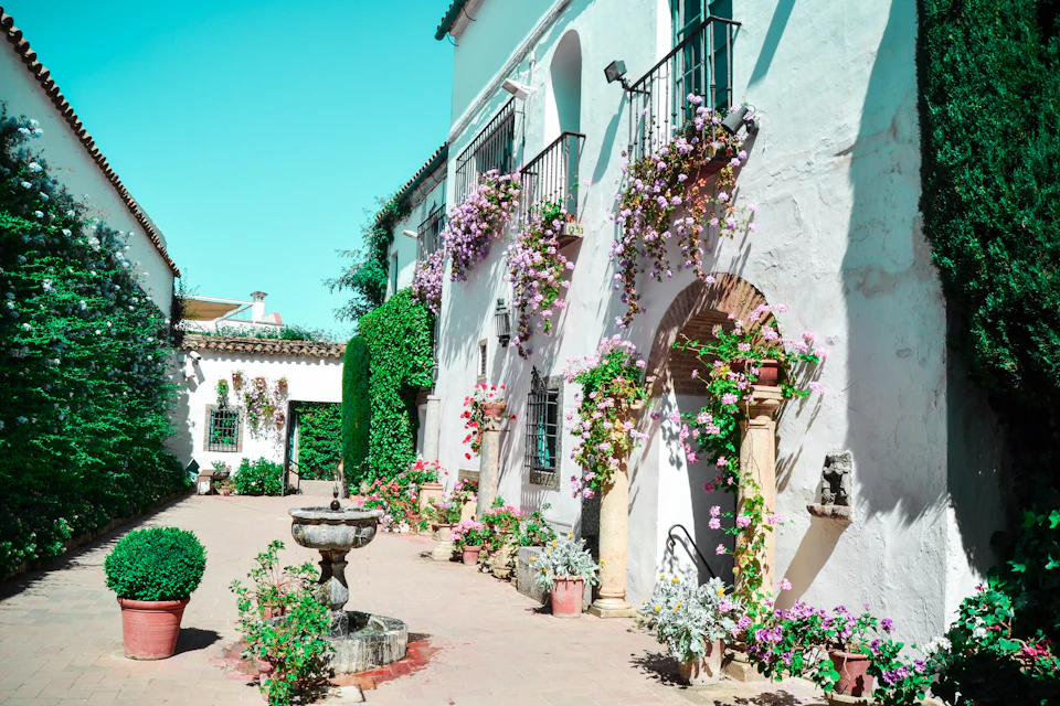 Courtyard with white walls, archways, and balconies adorned with flowering plants. Central stone fountain and potted plants.