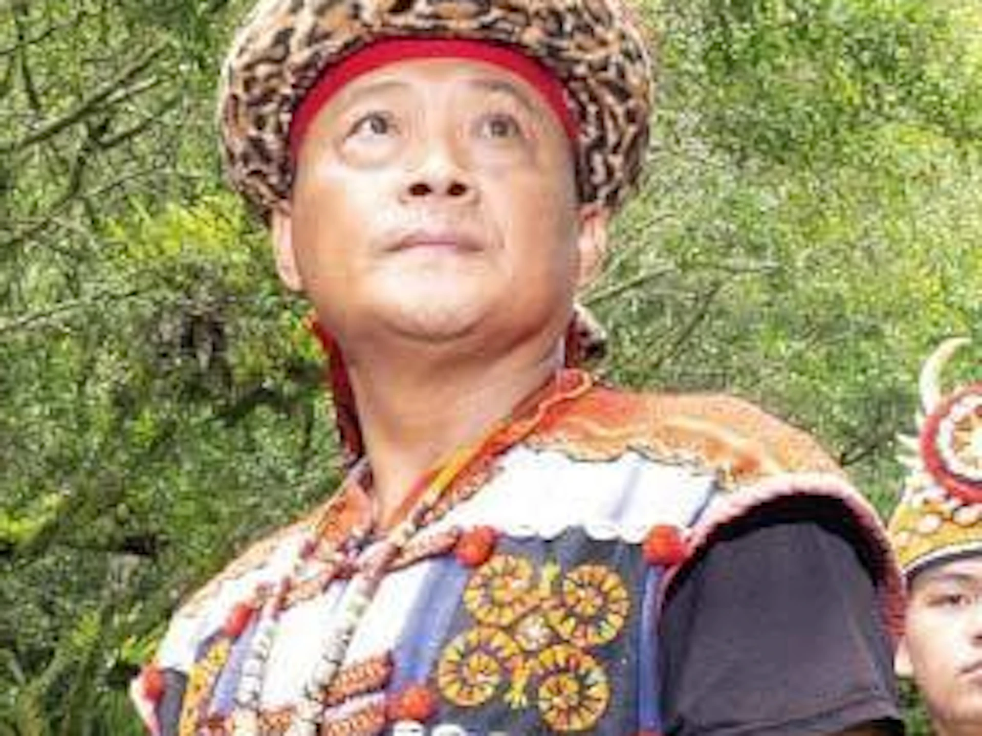 A group of people in traditional colorful attire with headdresses stand outdoors on a stone pavement surrounded by trees.
