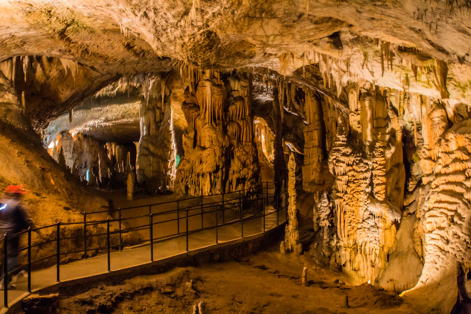 An illuminated cave with stalactites and stalagmites, featuring a pathway with a black railing.