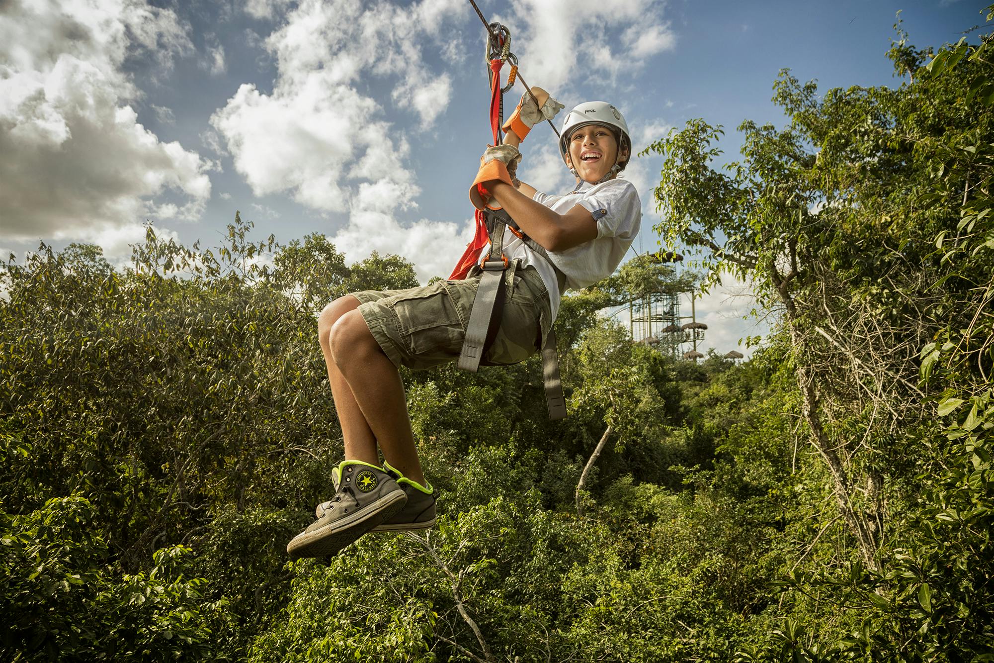 ヘルメットとハーネスを着用している人が、バックグラウンドでプラットフォームを見ながら、森の中をジップアップしています。