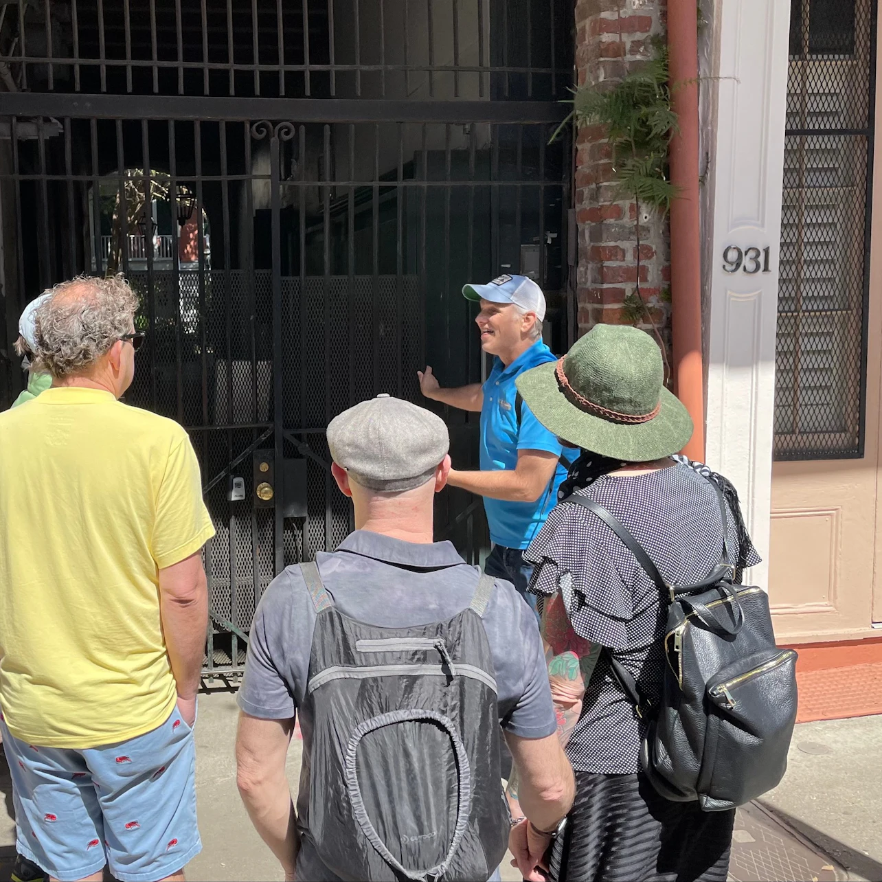 A group of people stands near a building entrance, with one person gesturing towards the gated doorway, in a sunny urban setting.