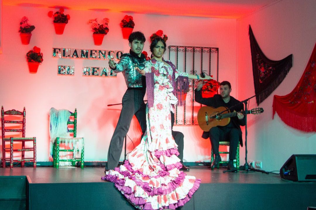 Na scenie występują dwie tancerki flamenco w stroju tradycyjnym, w towarzystwie siedzącego w tle gitarzysty.