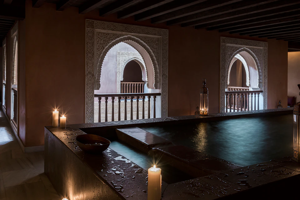 Dimly lit pool area with candles and ornate arched windows featuring intricate designs and wooden railings.