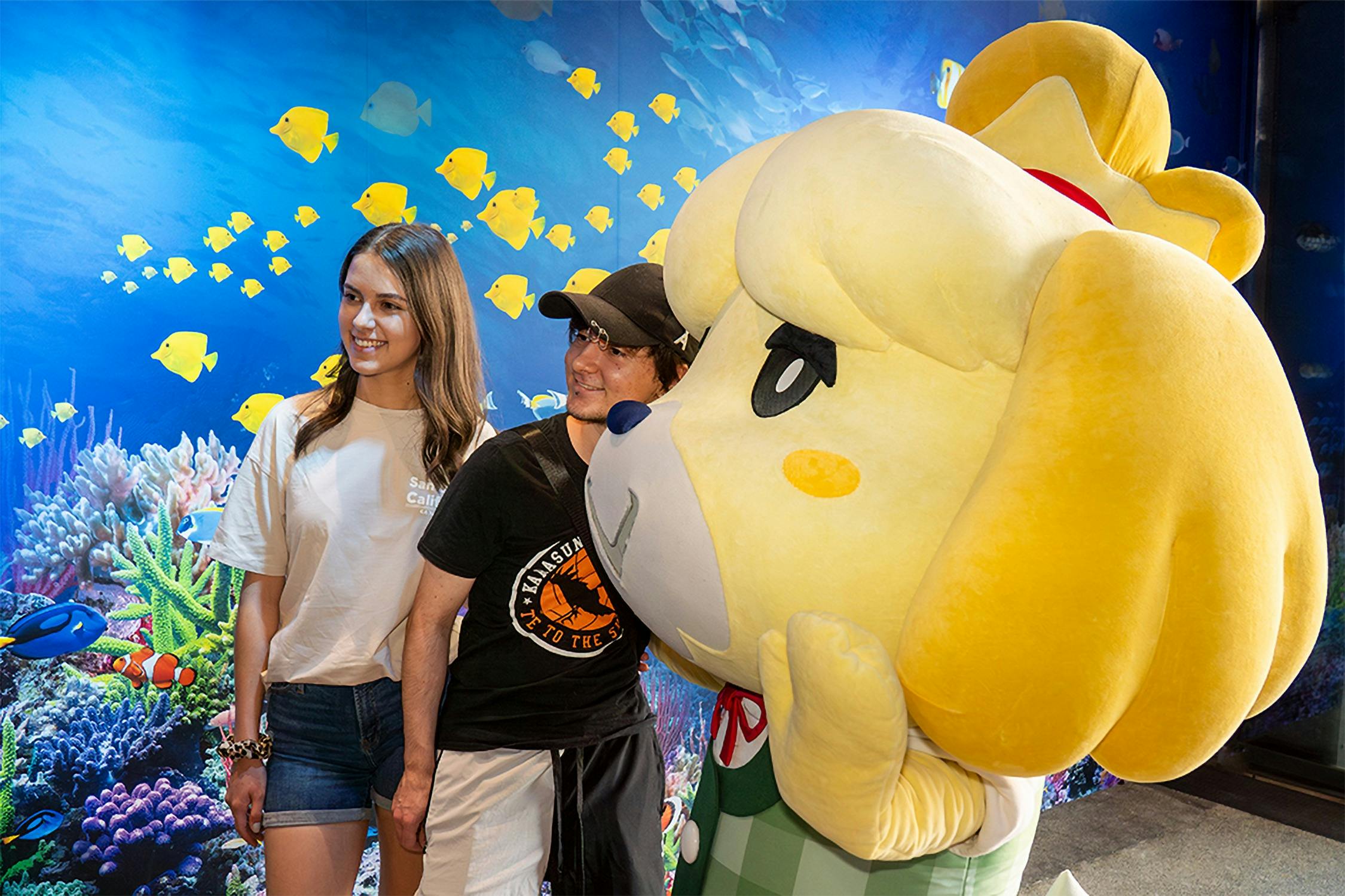 Dues persones parades al costat d'un gran personatge de felpa amb un fons de temàtica submarina amb peixos i corall.