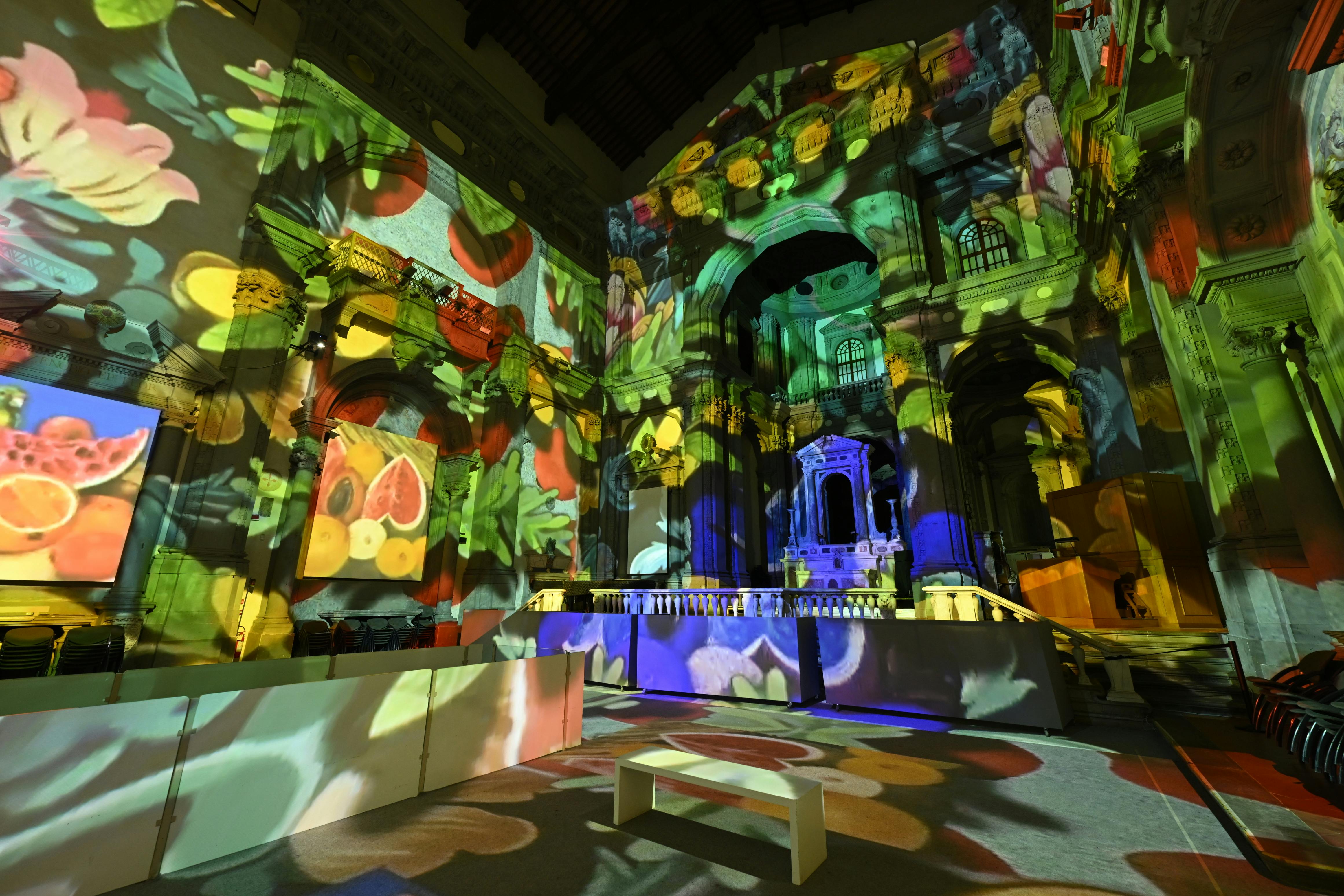 A church interior illuminated by colorful fruit-themed light projections, with a white bench in the foreground.