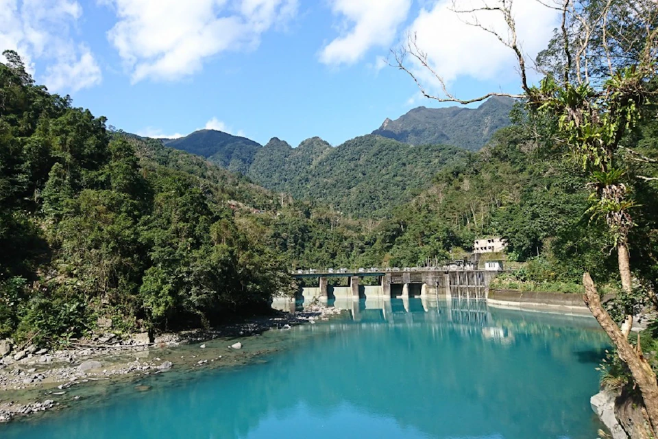 A serene landscape with a dam across a turquoise river, surrounded by forested hills under a partly cloudy sky.