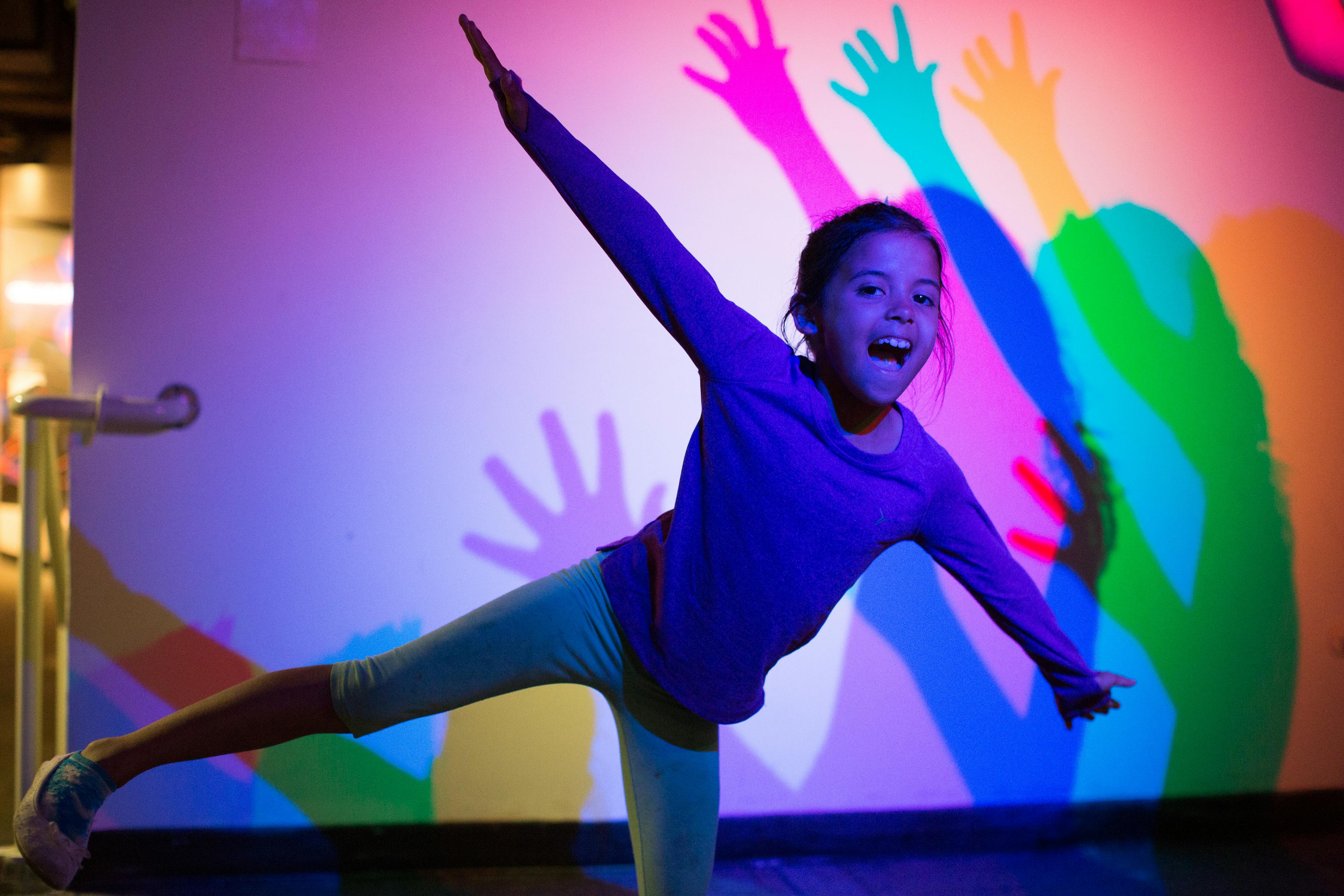 Ένα κορίτσι με μοβ μπλούζα ποζάρει παιχνιδιάρικα μπροστά από έναν πολύχρωμο τοίχο με πολλαπλές επικαλυπτόμενες σκιές.