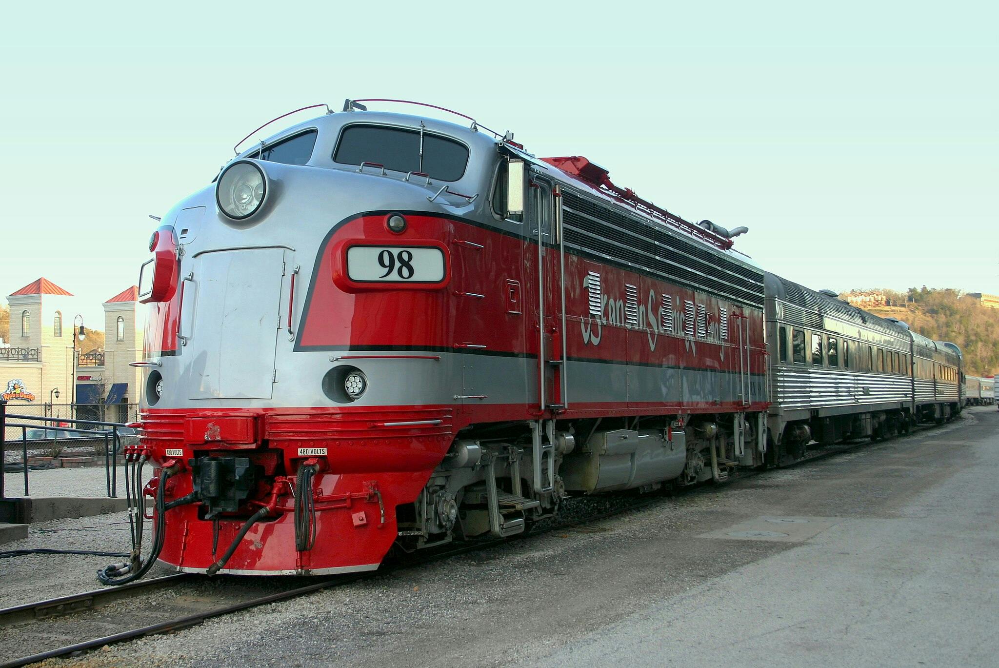 A vintage red and silver locomotive labeled "98" is parked on train tracks near buildings with red-roofed towers.