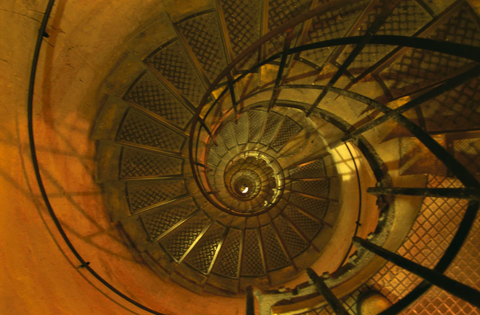 A spiral staircase with metal steps and railings, viewed from above, creating a circular pattern leading downwards.