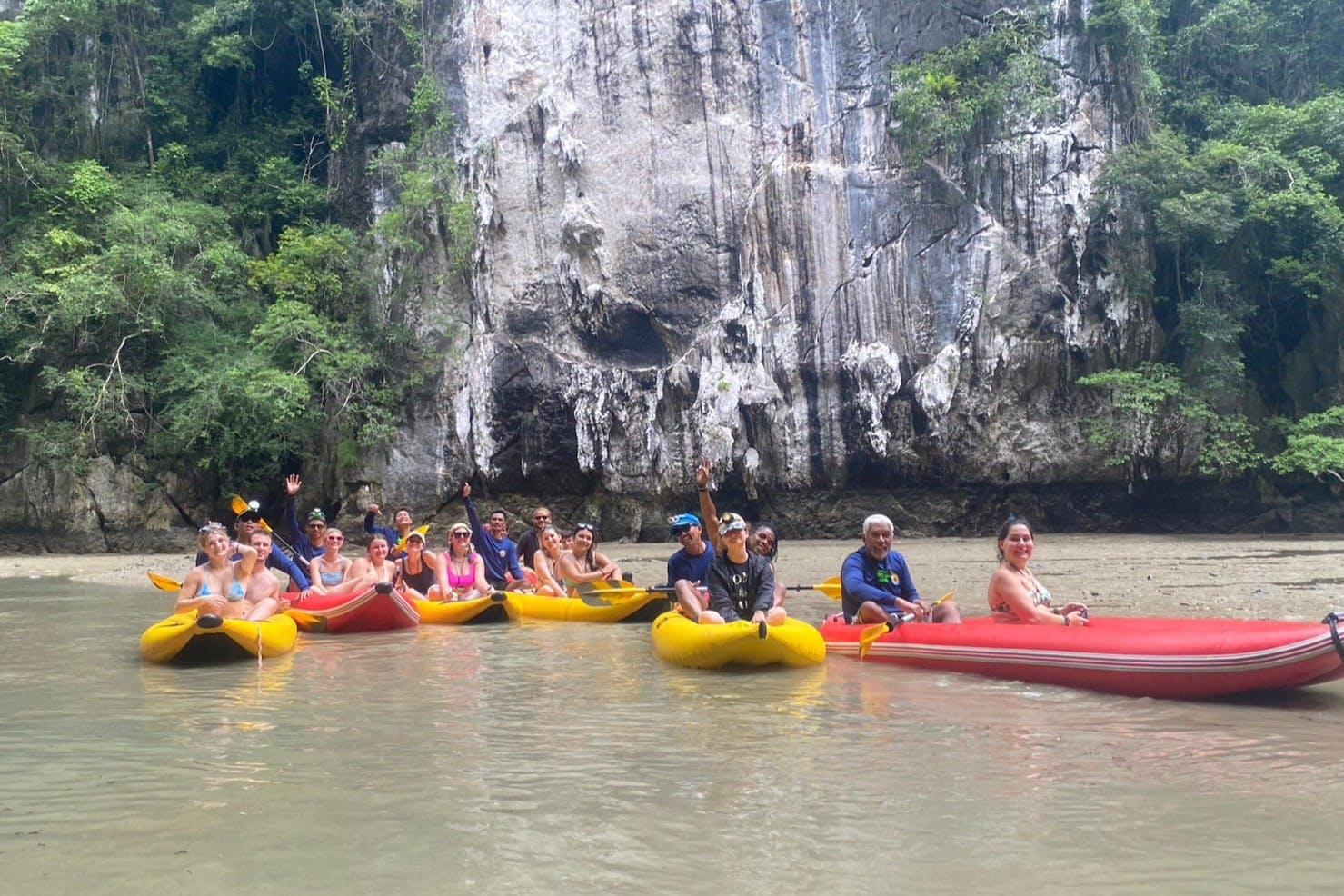 Lidé ve žlutých a červených kajakech pádlují na řece s velkým skalnatým útesem a hustou zelení v pozadí.